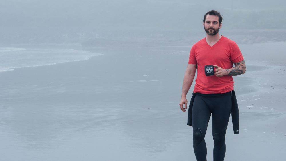 A man walking along a beach drinking Cooper's Cask Coffee and enjoying the coffee flavor profile