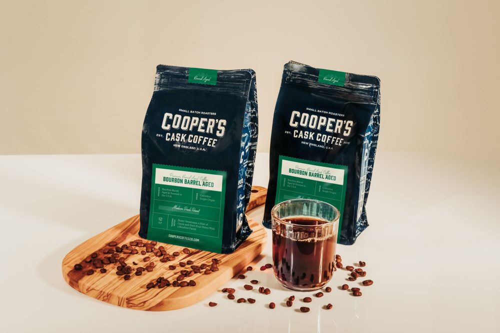 cooper's cask coffee bourbon coffee beans with cutting board, coffee cup, and whole bean coffee sprinkled on the counter