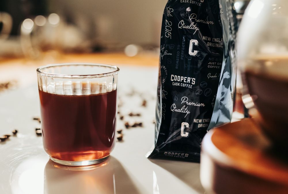 a cup of coffee sitting next to a bag of beans, with whole bean coffee sprinkled on the counter