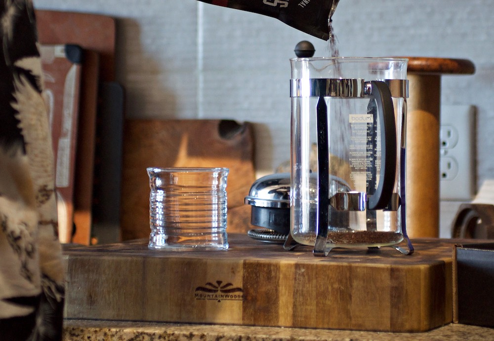 A french press beaker sits on a wooden block in a kitchen as a man pours coffee into it as one of the coffee brew methods for gourmet coffee