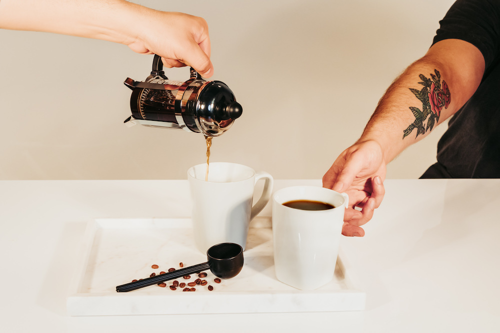 one hand pours french press bourbon infused coffee into white cup with beige background