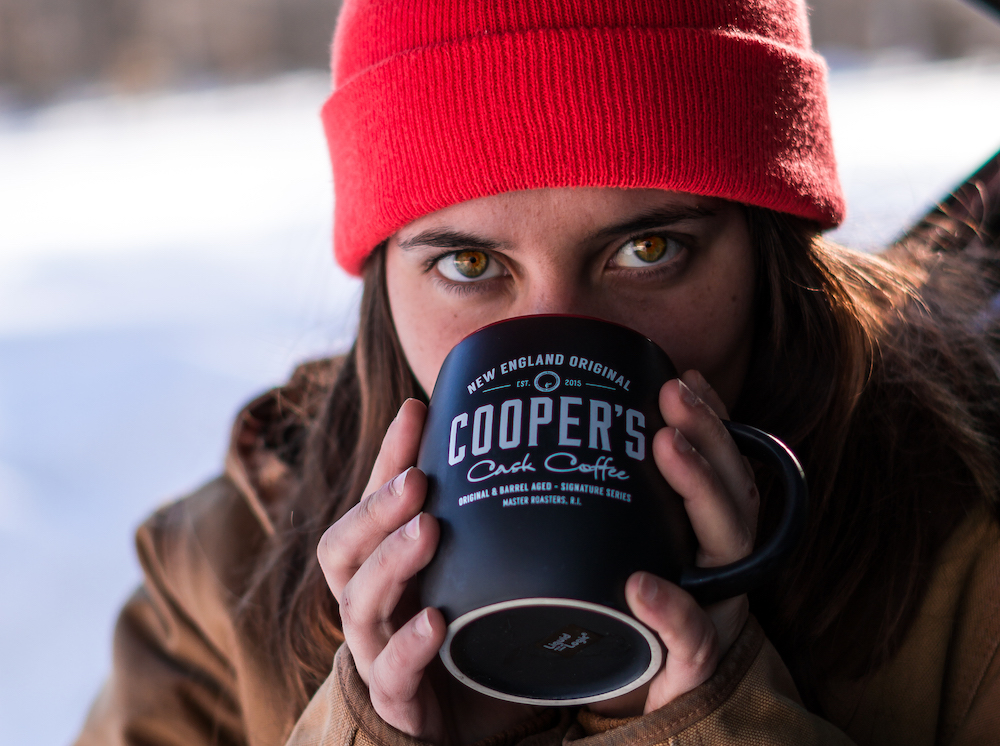 A woman out camping in a coat & hat drinks bourbon coffee from a Cooper's cup to show outdoor coffee brew methods