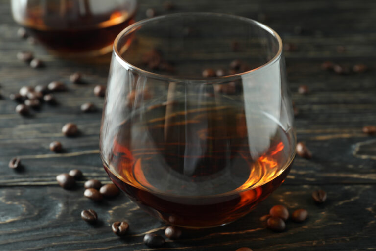 a snifter of bourbon-flavored coffee on a dark wood surface with coffee beans around it to represent bourbon infused coffee